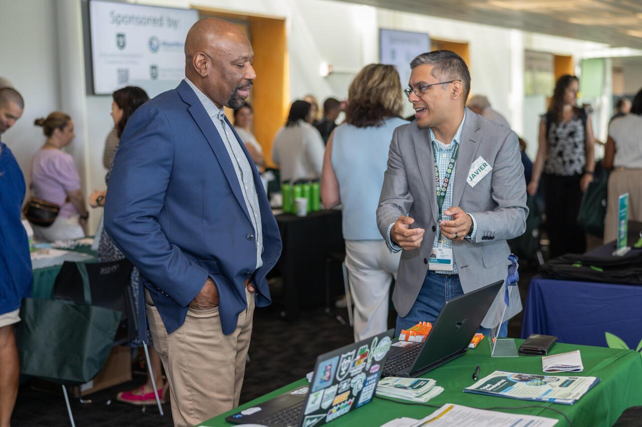Two men in blazers talk animatedly at a green table at a professional event.