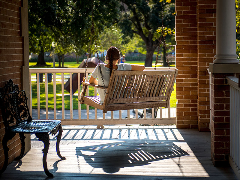 A young woman sits on a swing on Cudd Hall. Her back is to the camera and she appears to be reading a book.